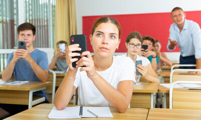 Interested girl high school student using mobile phone to find necessary information on lecture in class. Concept of using technology in education