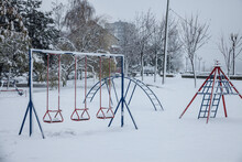 Old Children Playground, Slides And Swings, In A Park Covered In Snow In Serbia, In Eastern Europe, During A Freezing Dark Evening Winter Afternoon With Residential Buildings In Background...