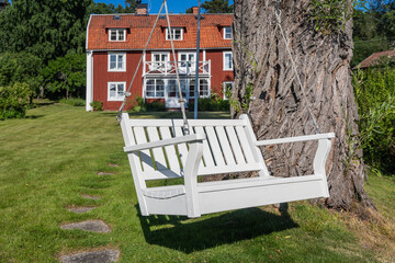 Wall Mural - A white wooden garden swing hanging on a tree. Classical swinging bench on the green lawn and trees surrounded. Landscape design: beautiful flowers on foreground. Red traditional cabin in background.