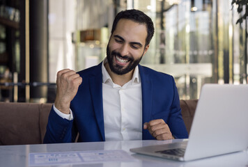 Wall Mural - Overjoyed  Moroccan businessman using laptop computer working online sitting in modern office. Happy trader receive payment celebration success at workplace