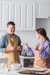 © LIGHTFIELD STUDIOS - Positive teenager with down syndrome holding smartphone near friend with dough in kitchen.