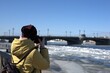 A man photographs the river and the bridge. Ice drift on the river. Spring weather in the city. Warm jacket and hat. Blagoveshchensky bridge across the Neva in St. Petersburg.
