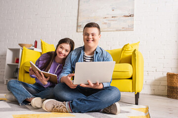 Positive teenagers with down syndrome holding laptop and notebook on floor at home.