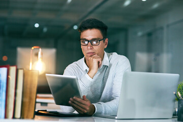 Leinwandbilder - He sure is dedicated to the job. Shot of a handsome young businessman using a digital tablet in an office at night.