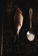 Fish with red eyes and fins next to spoon with salt on black surface of table