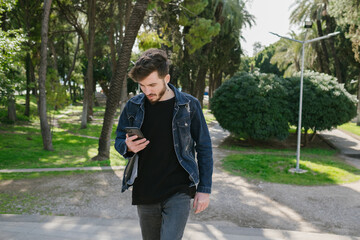 Wall Mural - Young man wearing jeans surfing on phone or reading message while walking on city park. Man typing a message to girlfriend.