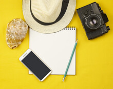 Vacation And Travel. A Notepad On A Bright Yellow Background, A Straw Hat, A Shell, A Camera And A Phone. Summer Background With Notebook For Notes And Space For Copy.