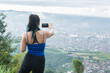 latina woman on the top of a mountain shooting a video of the city of Pereira-Colombia, with her mobile phone in horizoltal. girl hiking in the colombian mountains.