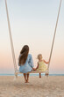 © somemeans - Young mother and little blond girl sit on swing on the beach during sunset. Mom and daughter on sea, magic sunset lights.