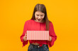 Stunned and surprised young girl looks into an open gift box. Portrait of teenager with gift in hands. Yellow background