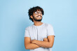 © Kateryna - Handsome attractive indian or arabian curly-haired guy, wearing t-shirt and glasses, standing over isolated blue background with crossed arms, looking away, smiling happily