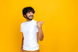 Positive curly haired indian or arabian millennial guy, looking and pointing finger to the side to an empty mock-up space, while standing over isolated orange background, smiling happily