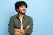 © Kateryna - Handsome charismatic successful indian or arabian curly-haired guy in stylish clothes, standing with crossed arms over isolated blue background, looking away, smiling positively