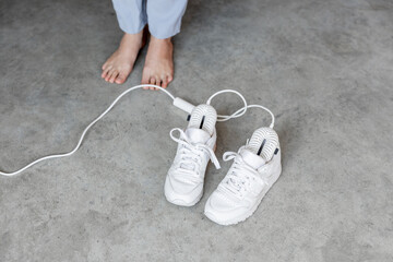 Woman stands barefoot near white sneakers drying with electric shoe dryer on the floor. Concept of modern technology and smart home gadgets