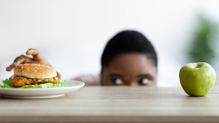 Overweight black woman looking at hamburger from under table, selecting between healthy and unhealthy food at home