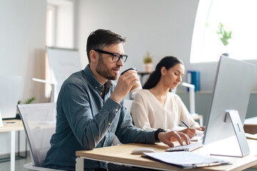 Canvas Print - Handsome young man using computer, drinking coffee, working with his female colleague at open space office