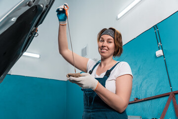 Wall Mural - Portrait of young woman mechanic in overall and gloves repairs oil dipstick of car. Bottom view. The concept of women's equality