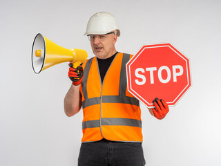 Man in road worker's uniform. Builder with loudspeaker. Road worker shows stop sign. Portrait of road repairman with loudspeaker. Builder shouts into megaphone. Man builder on light background