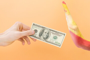 Say no dollar. The hand gesture of Spain rejects dollars. One arm holds 100 dollars, and the other arm painted with the Spanish flag doesn't want to take it. Isolated on yellow background. Wrong