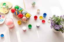 On A White Table, In Daylight, There Are Brushes In A Glass Of Water, Multi-colored Eggs In A Tray, Jars Of Paint.