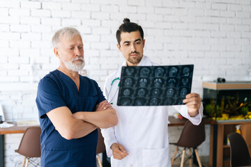 Wall Mural - Portrait of mature older surgeon and young physician doctor standing and discussing together history disease of patient using MRI scan of head. Concept of modern medicine and health care.