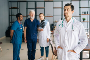 Wall Mural - Portrait of thoughtful young male doctor wearing white uniform standing in medical meeting office, looking away. Multi-ethnic team of physician working in background, discussing xray scan of patient.