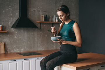 Wall Mural - Sporty woman sitting in the kitchen with a bowl and a spoon, eating. Vegetarian healthy food. Healthy lifestyle. Healthy eating, dieting, weight.