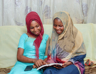 Wall Mural - Two happy African Nigerian female Muslim sisters or friends with Hijab scarf, sitting together while studying with a book placed on their laps