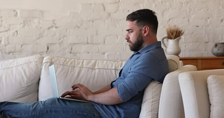 Concentrated young man use modern tech in freelance work type on laptop keyboard sit on couch at home. Thoughtful millennial guy software developer create program code remotely think on new solution