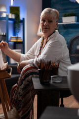 Portrait of gray haired older hobby artist studying shading light on still life using model vase in home studio. Retired woman drawing with hb pencil on canvas in front of easel while sitting down.