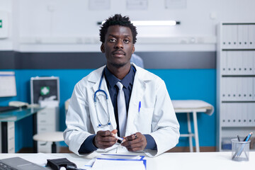 Wall Mural - African american polyclinic general practitioner expert sitting in modern clinic cabinet wearing stethoscope. Hospital physician specialist at work sitting at desk in medical workspace.