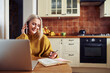 © baranq - Smiling mature woman sitting in the kitchen at home talking on the phone using laptop writing in notebook