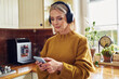 © baranq - Beautiful mature woman using mobile phone listening music on wireless headphones in kitchen