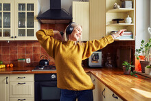 Joyful Mid Adult Woman Dancing In Kitchen Listening Music On Wireless Headphones