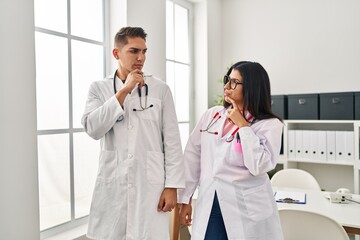 Poster - Young doctors wearing uniform and stethoscope at the clinic thinking concentrated about doubt with finger on chin and looking up wondering