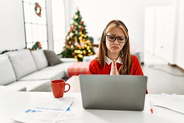 Wall Mural - Young caucasian girl sitting on the table working using laptop by christmas tree begging and praying with hands together with hope expression on face very emotional and worried. begging.