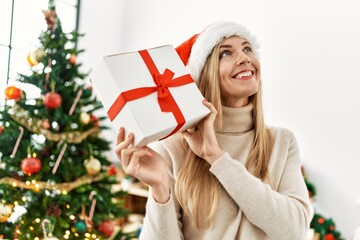 Wall Mural - Young blonde woman holding gift standing by christmas tree at home