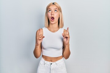 Poster - Beautiful blonde woman wearing casual style with sleeveless shirt amazed and surprised looking up and pointing with fingers and raised arms.