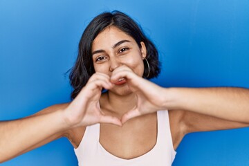 Sticker - Young hispanic woman standing over blue background smiling in love doing heart symbol shape with hands. romantic concept.