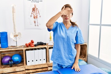 Canvas Print - Middle age hispanic physiotherapist woman working at pain recovery clinic covering eyes with hand, looking serious and sad. sightless, hiding and rejection concept