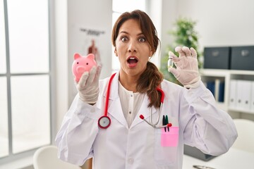 Canvas Print - Hispanic doctor woman holding piggy bank afraid and shocked with surprise and amazed expression, fear and excited face.