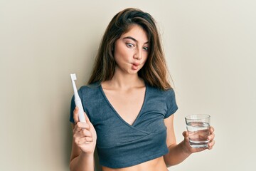 Canvas Print - Young caucasian woman holding electric toothbrush and water glass making fish face with mouth and squinting eyes, crazy and comical.