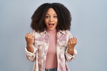 Poster - Young african american woman standing over blue background celebrating surprised and amazed for success with arms raised and open eyes. winner concept.