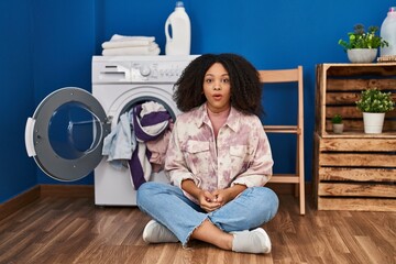 Sticker - Young african american woman sitting on the floor doing laundry scared and amazed with open mouth for surprise, disbelief face