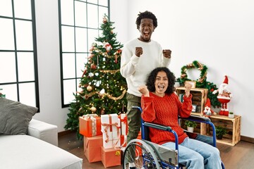 Poster - Young interracial couple with woman sitting on wheelchair by christmas tree celebrating surprised and amazed for success with arms raised and open eyes. winner concept.