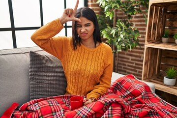 Sticker - Young hispanic woman sitting on the sofa drinking a coffee at home making fun of people with fingers on forehead doing loser gesture mocking and insulting.