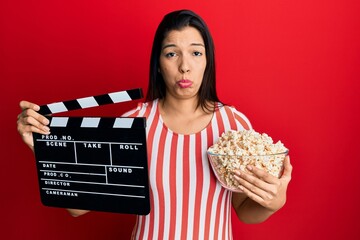 Poster - Young latin woman holding video film clapboard and popcorn depressed and worry for distress, crying angry and afraid. sad expression.