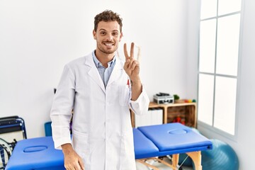 Wall Mural - Handsome young man working at pain recovery clinic smiling looking to the camera showing fingers doing victory sign. number two.