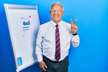 Wall Mural - Senior man with grey hair standing by business blackboard smiling with happy face winking at the camera doing victory sign with fingers. number two.