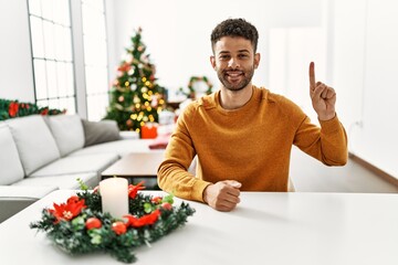 Sticker - Arab young man sitting on the table by christmas tree showing and pointing up with finger number one while smiling confident and happy.